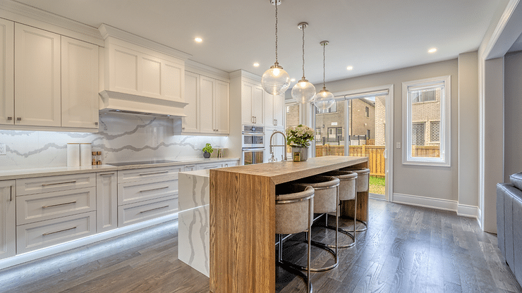 Modern kitchen with white cabinets, marble backsplash, wooden island, and stools. Pendant lights hang above. Large windows with a garden view.